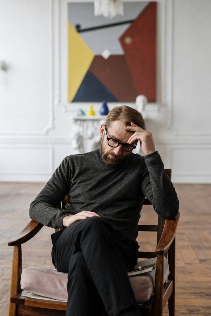 pexels-photo-4100670-4100670 A man sits pensively in a chair, surrounded by abstract art in a stylish room.